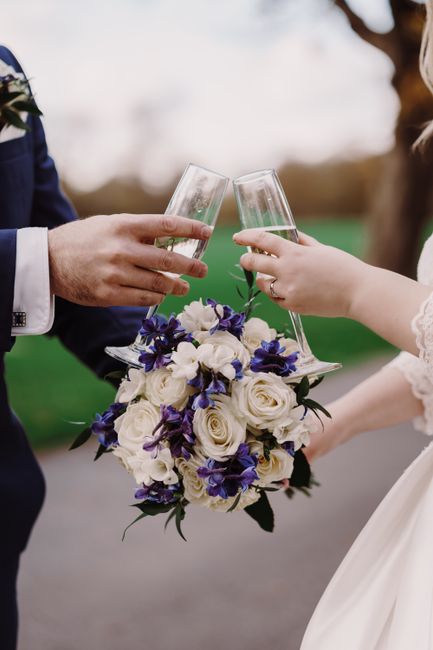 Close-up of a bride and groom holding champagne flutes in a celebratory toast, with the bride holding a floral bouquet comprising creamy white roses and deep purple irises, accented with green foliage