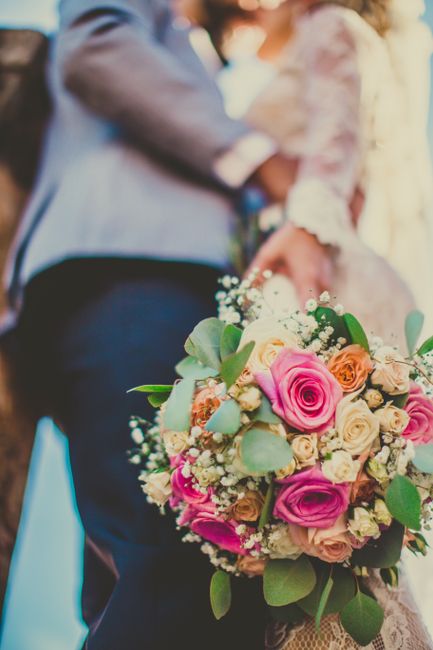 A vibrant bouquet of fresh flowers features pink roses, cream-colored roses, peach roses, and sprigs of baby's breath, accented with green eucalyptus leaves. The bouquet is held by a person with a lac