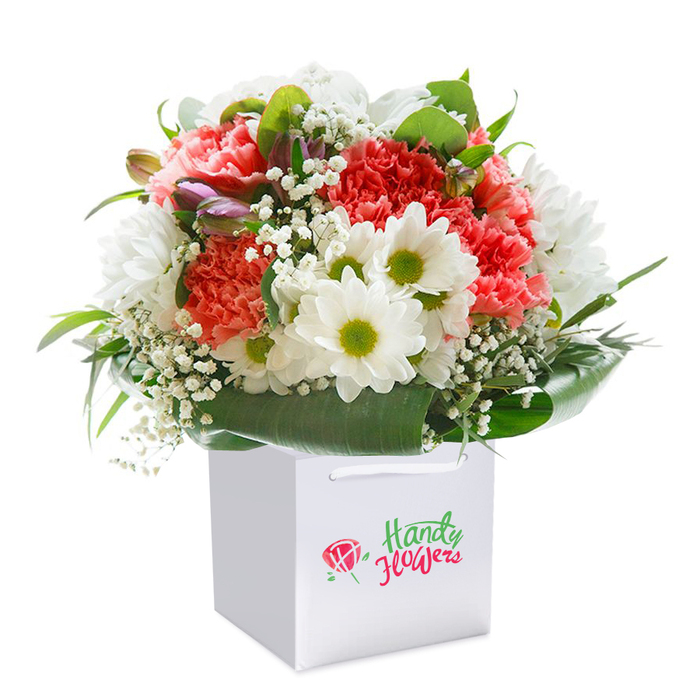 A lavish blend of colour and texture, this Norbiton-inspired bouquet is artfully presented in a contemporary, white square gift bag. At its heart, soft coral and peach carnations display feathery, ruffled petals, their vibrant tones radiating energy-much like the lively atmosphere along Norbiton's Coombe Road. Alternating with them, crisp white daisies show off charts of yellow-green at their centres, infusing the ensemble with a cheerful brightness that recalls the flowers found blooming near Kingston University. Scattered throughout are airy bursts of white gypsophila, their dainty blossoms imparting a dreamy, delicate feel that quietly anchors the bouquet. On the left, slender purple-tinged buds emerge, suggesting new beginnings and adding depth. The arrangement is enveloped by generous, glossy deep green leaves that arc around the base, forming an elegant frame reminiscent of the greenery that marks Norbiton's tree-lined avenues. Each bloom is expertly placed, creating a plush, rounded silhouette that celebrates floristry at its finest. The sturdy white bag features ribbon handles that peek from the top, crafted for convenient gifting as you wander through the borough. Set against a stark white backdrop, the flowers stand out in vivid detail, their freshness and quality clear. This vibrant bouquet is perfect for same-day delivery in Norbiton-an ideal choice for birthdays, anniversaries, or simply to make someone's day brighter in this close-knit corner of southwest London.