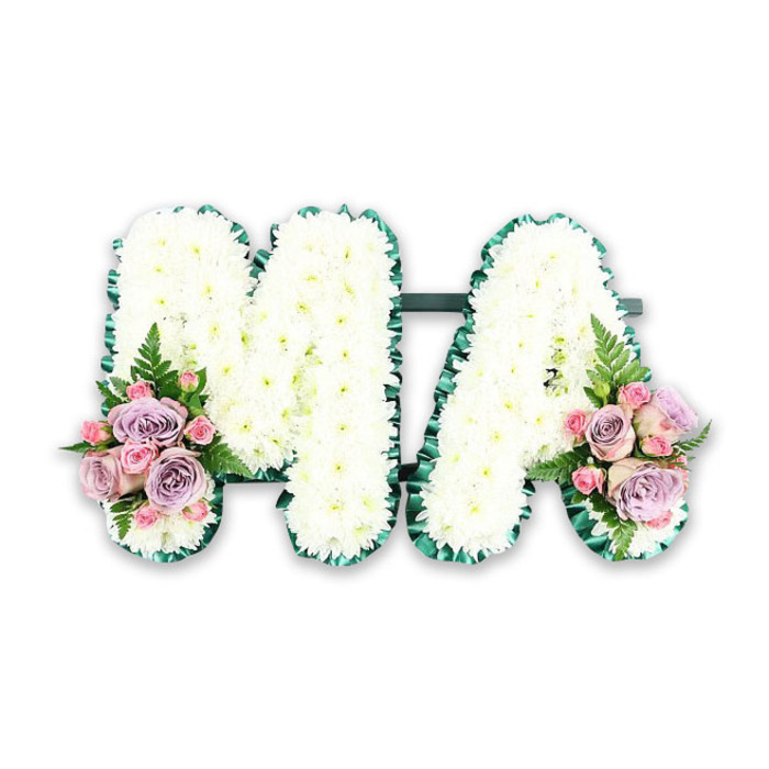 A personalised funeral flower arrangement spells out the word MA in bold capital letters, conveying the deep bond so often felt for a mother figure in families across Norbiton. Each letter is meticulously covered in compact, snow-white chrysanthemums, the blossoms arranged in tight formation to create a smooth, almost quilted texture. Their petals appear soft and feathery, catching the light with a gentle, matte glow that enhances the sense of stillness and peace. Tracing the outline of both letters is a band of rich, emerald green satin ribbon, gathered and pleated with precision. This ribbon frame creates a clear, defined silhouette, its slight sheen and darker tone setting off the white blooms in a way that feels both classic and contemporary. Subtle touches of greenery are tucked just beneath, adding a whisper of natural depth. At the bottom left point of the M and the bottom right of the A, matching floral posies introduce a romantic, pastel palette. Here, lilac and blush-pink roses unfold in layered petals, joined by smaller pink spray roses that add texture and a sense of gentle abundance. Between the roses, light, airy fern fronds curve outward, their soft green frills offering movement and echoing the relaxed greenery you might see in the gardens around Norbiton and Kingston. The entire tribute is photographed on a clean, white background, ensuring that the focus remains firmly on the craftsmanship and symbolism. It is the kind of bespoke letter tribute a family might choose for a crematorium or church service, expressing gratitude, love and lasting remembrance for a beloved Ma, Mum or Mama whose presence shaped the heart of the home.