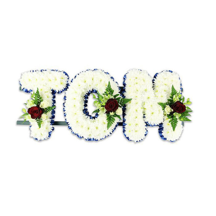 A solemn and beautifully crafted memorial arrangement reads TOM in large, sculpted letters formed entirely from tightly clustered white chrysanthemums, their small heads packed to create a pillowy, tactile surface that reflects soft, natural light. Each letter is edged with a vivid royal blue ribbon that traces its outline with deliberate precision, providing a crisp, dignified frame against the pure white florals. Within the center or lower area of each character is a compact spray built around a single, deep red rose - the visual and emotional anchor - accompanied by smaller creamy white spray roses and graceful fern fronds which unfurl into verdant accents. The interplay of velvet red, ivory and cool green against the white streamlines the composition and fosters a sense of reverence and warmth. Visible beneath the letters is a dark green or black metal support bar, a simple but necessary structural element that holds the tribute upright when set on a chapel easel or beside a graveside marker. The quiet geometry of the design, the careful placement of each accent spray, and the choice of materials reflect the hands of experienced florists accustomed to local ceremonies at Norbiton Park chapels and nearby community centres. There is a subtle fragrance - the faint earth of chrysanthemum with a sweeter undernote from the roses - and an overall stillness in the piece that invites reflection. This bespoke name tribute is intended to offer consolation and a personalised gesture of remembrance for mourners and family.