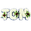The photograph captures a bespoke funeral flower arrangement spelling out TOM in striking, three-dimensional block capitals, the kind of named tribute often seen at services in Norbiton churches or local crematoria. Each letter is fully covered with pristine white chrysanthemums, arranged so tightly that the surface appears plush and gently domed, like a soft cushion of petals. Around the outer edges, a royal-blue satin ribbon runs in a continuous line, its smooth, shining texture neatly framing each letter and giving the piece a polished, respectful look. Carefully placed at key points along the name are small, artful sprays of accent flowers: deep crimson red roses that suggest love and devotion, dainty white or cream spray roses, and slender, elegant white blooms similar to freesia. These floral groups are surrounded by fresh green foliage and airy fern, adding a touch of natural movement and contrast against the uniform white chrysanthemum base. The letters sit side by side on a thin, dark metal bar that supports them in a straight horizontal row, ready to be positioned beside a coffin, on a hearse, or near the front of a memorial space. The plain white background in the image enhances the clarity of the design, allowing every detail-from the frilled ribbon to the clustered roses-to stand out. Overall, the tribute conveys a sense of quiet honour and lasting affection, a personalised farewell created for someone named Tom, reflective of the thoughtful sympathy designs that local Norbiton families often request when marking a final goodbye.