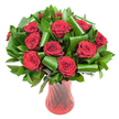 A lush, rounded bouquet of a dozen to fifteen deep crimson roses fills the frame, each bloom fully open with velvety petals spiralling from a tight centre to soft, unfurling edges. The wine-dark reds read intense and satiny under soft, even studio lighting that picks out the subtle veining on each petal and gives the heads a tactile, velvet appearance. The roses sit nestled in a generous collar of dark green foliage: flat, glossy leaves-suggestive of Ruscus or Salal-form a dense, verdant backdrop while broader, emerald Aspidistra-like leaves have been artfully rolled and curled into dramatic loops and cylinders. These sculpted greens create elegant architectural accents that cradle individual roses and add modern structure to the classic red arrangement. The stems are arranged in a tall, translucent ruby-red glass vase with a subtle ribbed or twisted surface that throws long vertical highlights, echoing the flowers' passionate hue and elongating the composition. Set against a crisp white background, the saturated reds and deep greens pop with striking clarity, the simple backdrop isolating texture and colour so the eye rests on form and detail. As a florist I imagine the soft, classic scent of red roses rising from the cluster-a familiar, comforting perfume that hints at anniversaries, Valentine's Day, or a romantic gift from Norbiton to Kingston. The overall impression is one of refined romance and deliberate design: traditional red roses given a contemporary twist by folded foliage and a sculptural vase, perfect as an elegant centrepiece or a heartfelt, memorable gesture.