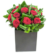 A modern, sculptural bouquet of deep crimson roses sits like a small, dramatic installation in a matte black cube, each bloom captured in a moment of velvety unfolding. The roses are rich and dense, petals spiralling outward in layered, velvet-soft whorls that catch the light and read as pure, romantic colour. Around them, broad glossy green leaves have been expertly looped and curled, their edges forming rhythmic arcs that both cradle the flowers and introduce a sense of movement - a deliberate, measured breeze sculpted into foliage. The square black vessel grounds the design with a clean, urban geometry; its matte surface absorbs light so the roses seem to glow from within. Lighting across the composition is bright and steady, highlighting the sheen on leaf surfaces and the plush texture of each petal, while casting gentle, minimal shadows that reinforce the bouquet's clarity. Close to the arrangement you might imagine a fresh, green top note - the crispness of washed leaves - gently overlaid by the classic, heady perfume of red rose, a scent that feels polished rather than cloying. This piece reads as contemporary romance: confident, composed, and meant to be both a visual statement and a tactile pleasure. It's equally at home brightening a sleek Norbiton office near the station as it is making a private anniversary feel significant. As a florist I see the balance of structure and softness here as intentional: a bouquet designed to make a memorable impression while remaining quietly elegant and undeniably tactile.